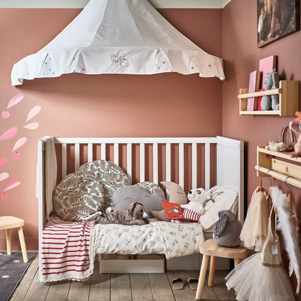 Ein Kinderzimmer mit Wänden in Rosa und einem weißen Babybett mit vielen kuscheligen Textilien und Stofftieren, einem weißen Betthimmel darüber und zwei Kinderhockern in Kiefer davor