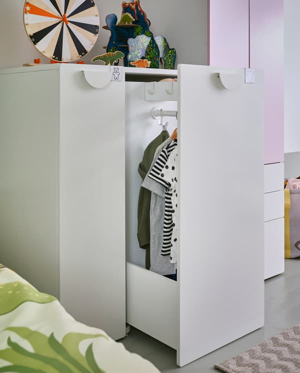 Ein Kleiderschrank mit Auszug in Weiß/Blassrosa, bei dem der Auszug offen steht. Darin befinden sich eine Kleiderstange und Haken. Daran hängt Kleidung an Kleiderbügeln.