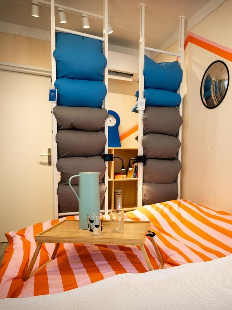 Ein kleiner Raum mit einem einem ELVARLI Raumteiler und einem Tablet auf dem sich ein Krug und Gläser befinden auf einem Bett.
