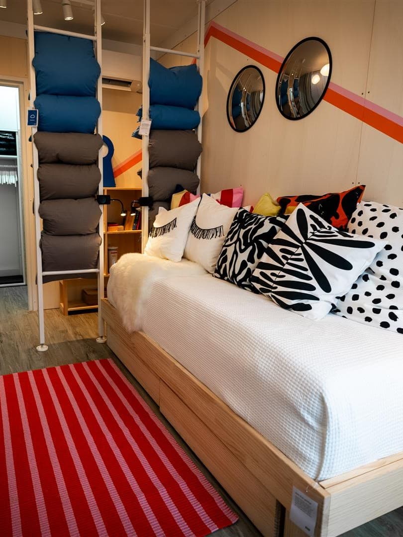 Ein kleiner Raum mit einem GLAMBERGET Bettgestell, vielen Kissen und einem ELVARLI Raumtrenner.