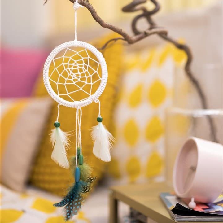 Ein kleiner weißer Traumfänger hängt neben einem Bett an einem Nachttisch.