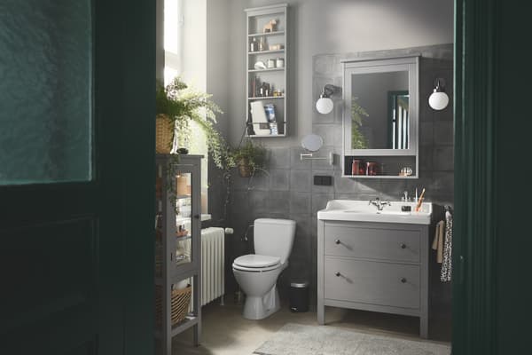 Ein kleines Badezimmer mit weißem HEMNES / RÄTTVIKEN Waschbeckenschrank, Spiegelschrank, Körben, Badematte und Wandleuchte.