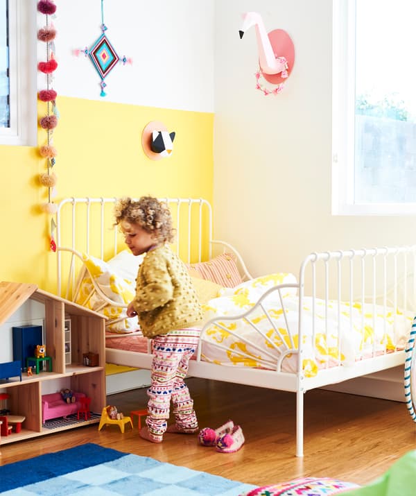 Ein kleines Kind in einem Kinderzimmer, u. a. mit MINNEN Bettgestell, ausziehbar, weiss. Daneben ist ein FLISAT Puppenhaus aus Holz zu sehen.