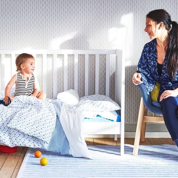 Ein Kleinkind sitzt in einem Babybett und blickt auf eine Frau, die daneben auf einem Stuhl sitzt.