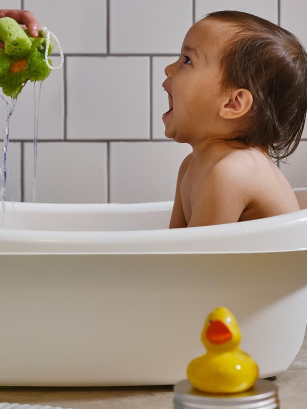 Ein Kleinkind sitzt in einer LÄTTSAM Babybadewanne und lacht, weil eine erwachsene Person Wasser aus einem grünen Schwamm spritzt.