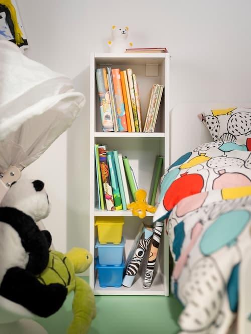 Ein KRAMIG Stoffspielzeug als Pandabär sitzt auf einer Stoffschildkröte neben einem weißen SMÅGÖRA Regal, das neben einem Kinderbett steht.