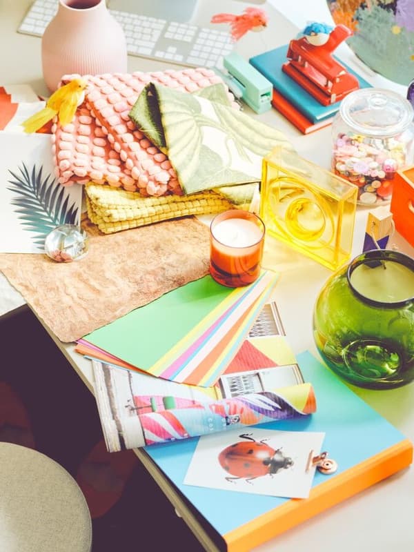 Ein kreativer und bunter Schreibtisch, bedeckt mit verschiedenen Materialien wie farbigem Papier, Stoffen und Magazinen. Es gibt auch Dekorationsobjekte wie eine Kerze, eine bunte Vase und einen Glasbehälter mit Süßigkeiten