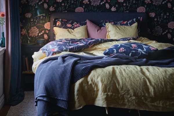 Ein kuscheliges Bett mit zwei Decken, Kissen und einer Tagesdecke. Auf dem Ablagetisch neben dem Bett sind eine Leuchte und ein Becher zu sehen.