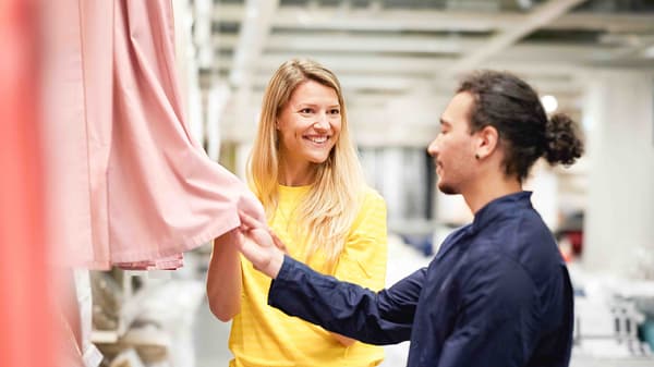 Ein lächelnder IKEA-Mitarbeiter berät eine Kundin bei der Auswahl von Vorhängen in einem Einrichtungshaus.