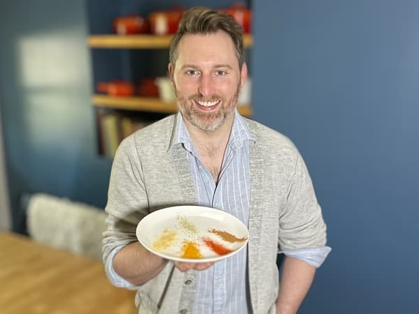 Ein lächelnder Mann hält einen Teller mit fünf verschiedenen Gewürzen in die Kamera.