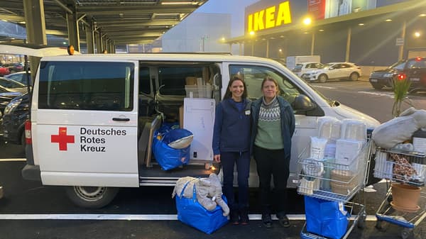 Ein Lieferwagen des Deutschen Roten Kreuzes steht auf dem Parkplatz vor einem IKEA Einrichtungshaus. Die seitliche Tür des Fahrzeugs ist geöffnet und zeigt geladene Sachspenden. Zwei Personen stehen davor, umgeben von blauen IKEA Taschen, Kartons und zwei beladenen Einkaufswagen mit Haushaltsartikeln und Textilien.