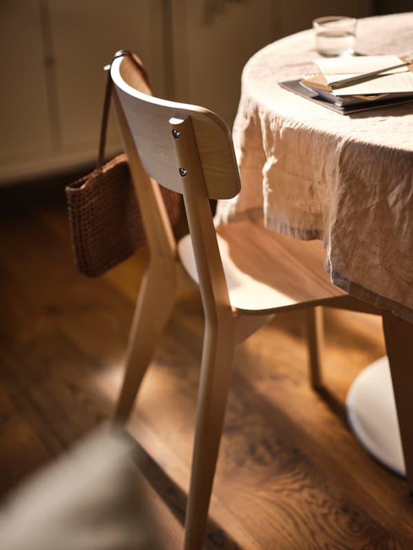 Ein LISABO Stuhl in Esche steht vor einem Tisch, auf dem eine Tischdecke liegt. An der Stuhllehne hängt eine Handtasche.