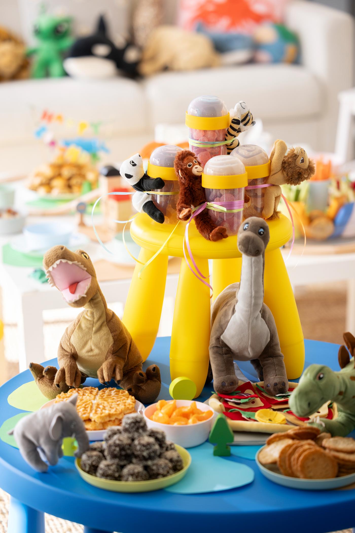 Ein MAMMUT Hocker steht als Etagere auf einem MAMMUT Tisch. Stofftierdinos bewachen die darauf angerichteten Snacks. 