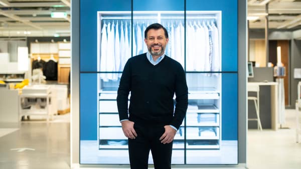 Ein Mann in dunkler Kleidung steht lächelnd vor einem großen digitalen Display, das ein leuchtend weißes Schranksystem mit hängender Kleidung zeigt. Die Szene befindet sich in einem IKEA-Einrichtungshaus.