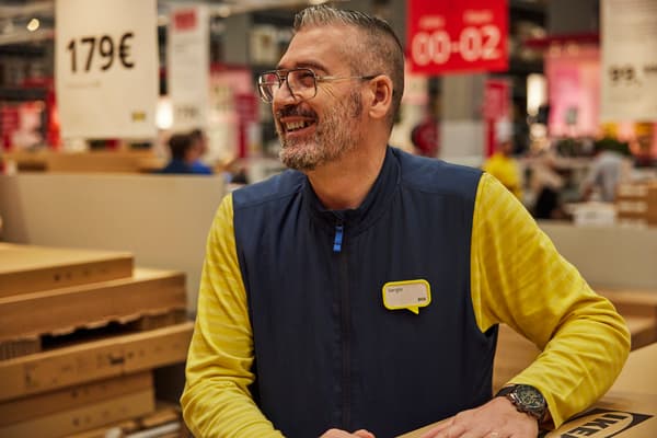 Ein Mann mit Brille, gelbem IKEA Shirt und blauer Weste lacht und stützt sich bei einem eingepackten IKEA Möbelstück ab.