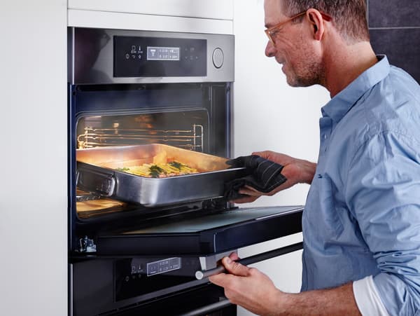 Ein mann öffnet einen Backofen mit Dampfgarfunktion. Es leuchtet und dampft aus dem Ofen. In dem Ofen befindet sich eine silberne Ofenform, in der ein Lachs mit Zitronenscheiben zubereitet wird. 
