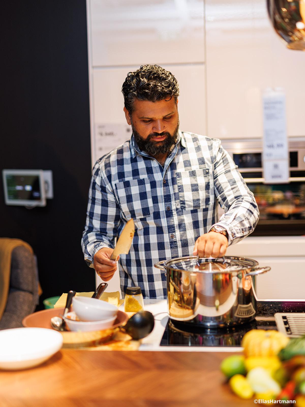 Ein Mann steht in der Küche am Herd und möchte den Deckel von einem Topf nehmen.