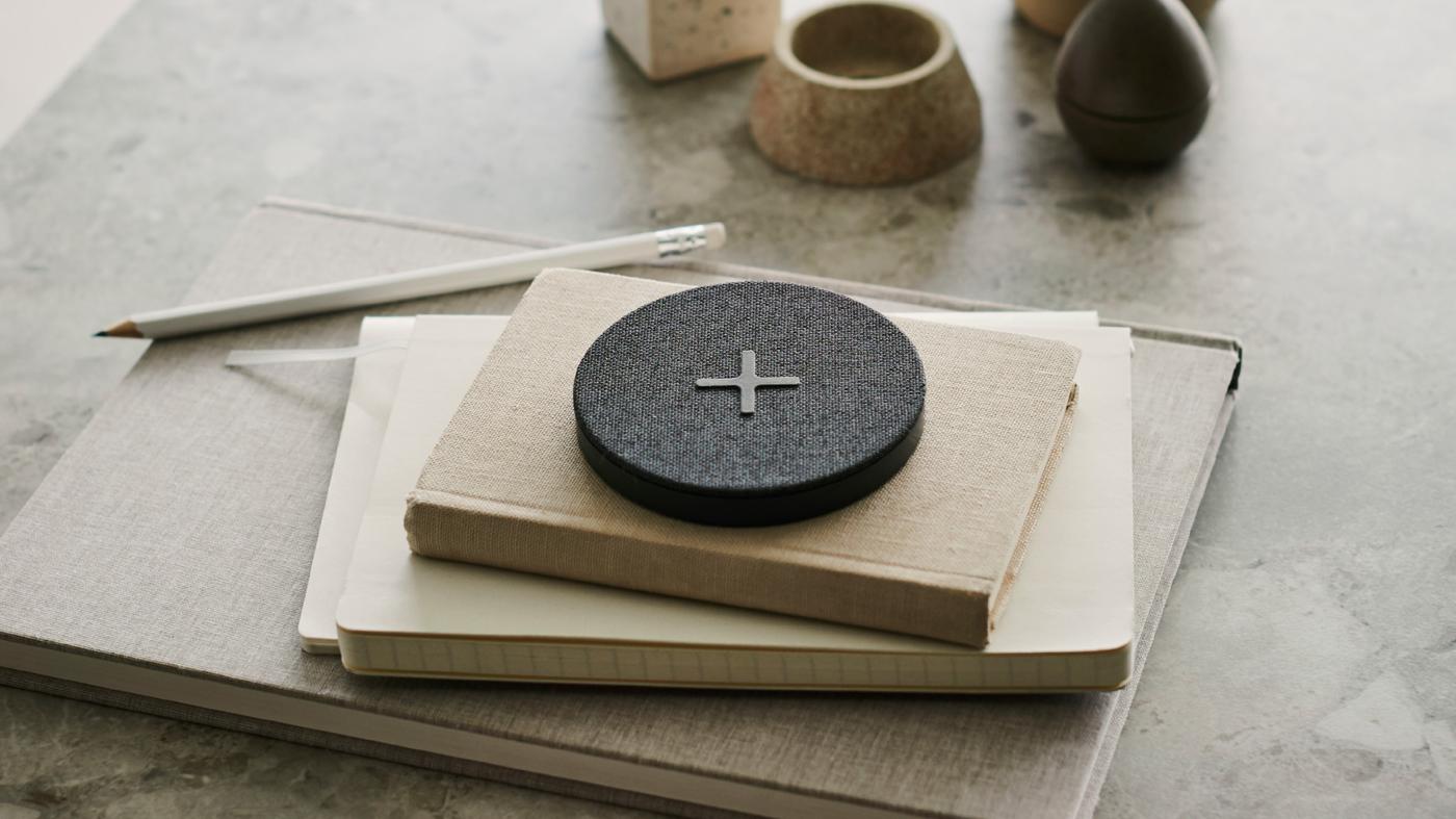 Ein NORDMÄRKE Ladepad mit einem großen Pluszeichen auf der grauen Textiloberfläche liegt auf einem Stapel Büchern.