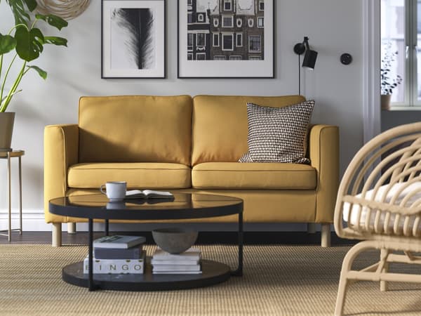 Ein ockerfarbenes PÄRUP 2er Sofa mit Holzbeinen steht vor einer grauen Wohnzimmerwand an der schwarz weiße Bilder hängen. Vor dem Sofa steht ein schwarzer runder Glas Couchtisch. 