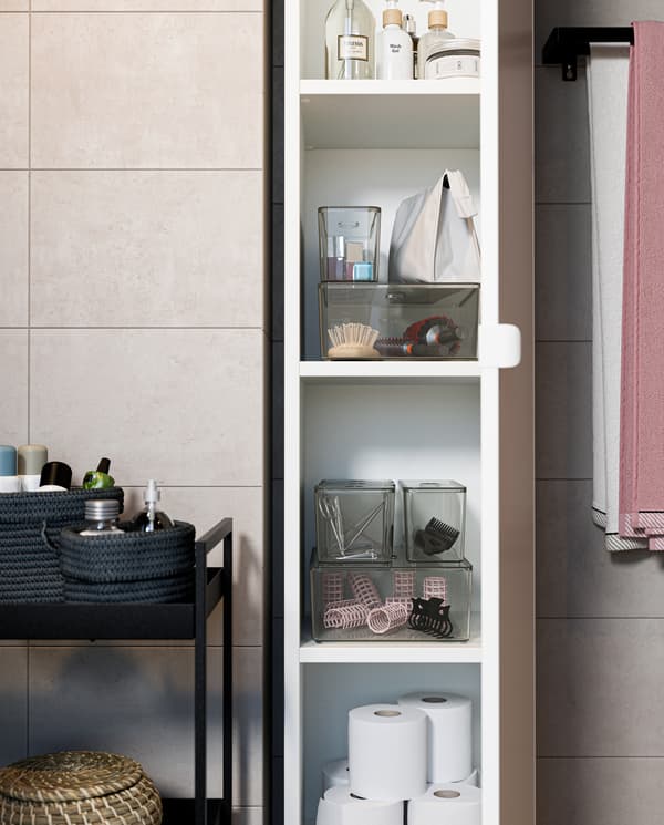 Ein offener Hochschrank in Weiß mit transparenten Kästen mit Deckel, Toilettenpapierrollen, Lotionen und vielem mehr. Daneben steht ein Servierwagen in Schwarz.