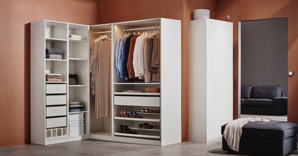 Ein offener, weisser Eckkleiderschrank und ein geschlossener, weisser Kleiderschrank vor einer rostbraunen Wand.