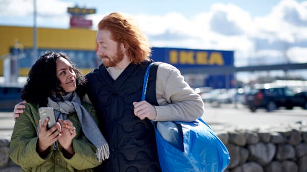 Ein Paar steht vor einem IKEA Einrichtungshaus. Die Frau zeigt dem Mann etwas auf dem Smartphone und er trägt eine gefüllte FRAKTA Tasche.