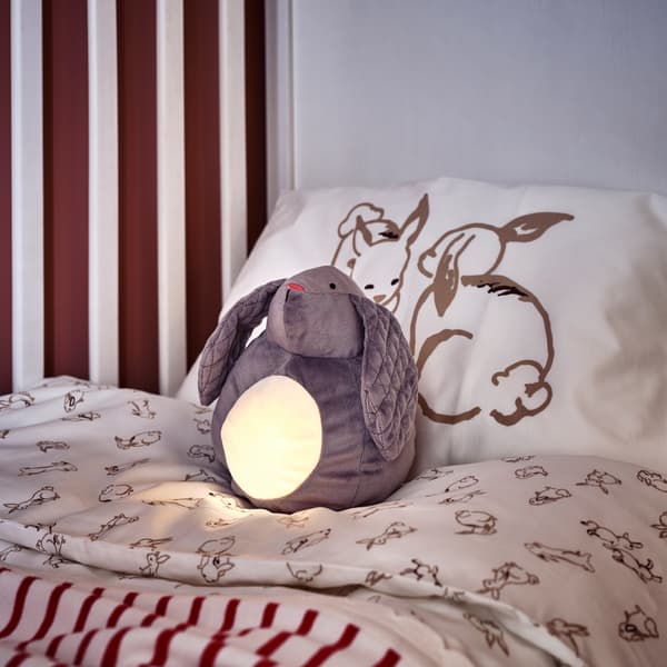 Ein PEKHULT Stofftier mit LED-Nachtlicht ist beleuchtet und liegt in einem Babybett mit Bettwäsche mit Kaninchenmuster in Weiß/Beige.