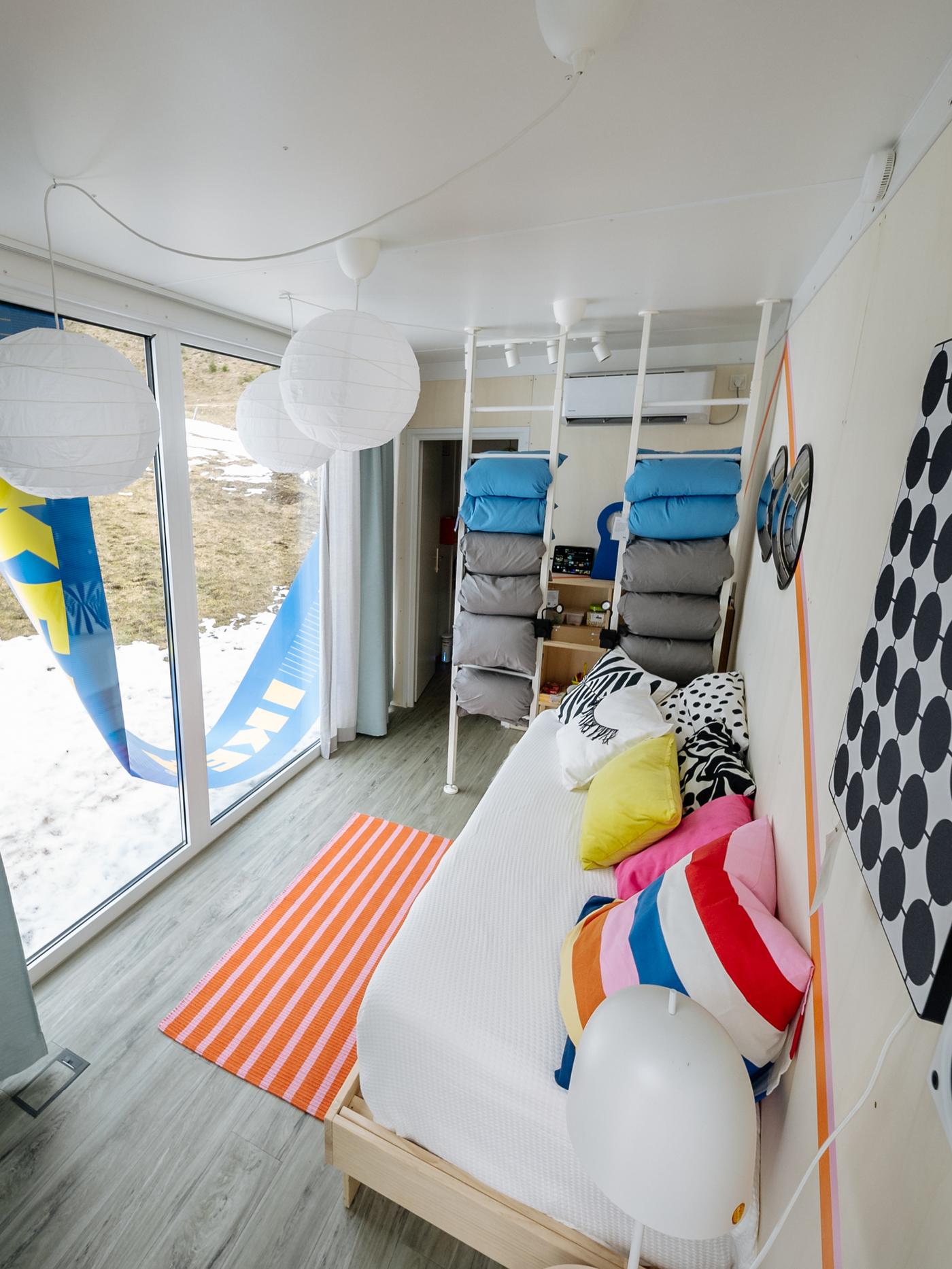 Ein Raum mit einem Bettsofa und Kästen. Es ist bunt eingerichtet mit boxen und Kissen und einem Orangenen Teppich.