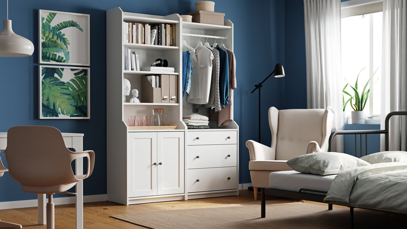 Ein Raum mit einer weißen HAUGA Kombination bestehend aus zwei Hochschränken, mit einem Schreibtisch, gerahmter Kunst, einem Ohrensessel vor dem Fenster und einem Bettsofa im Schlafmodus