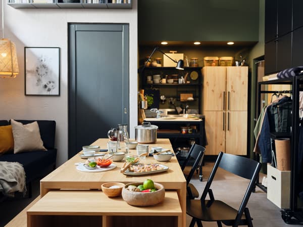Ein RÅVAROR Esstisch, ein RÅVAROR Regal mit Rollen und RÅVAROR Klappstühle stehen in der Mitte eines Apartments. Auf dem Tisch stehen Teller, Schüsseln und Gläser.