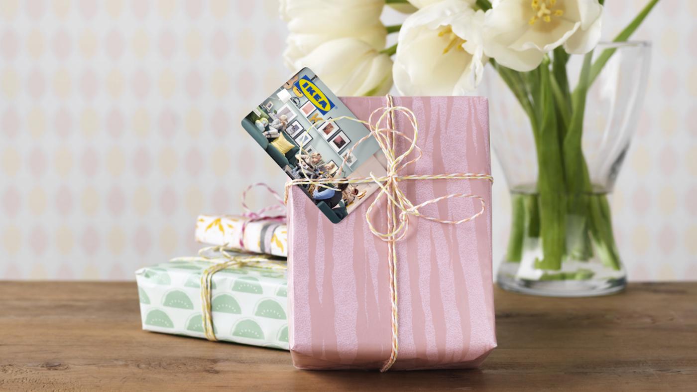 Ein rosa eingepacktes Geschenk, dekoriert mit einer Schleife und einer IKEA Geschankkarte. Im Hintergrund sind Blumen und weitere Geschenke zu sehen. 