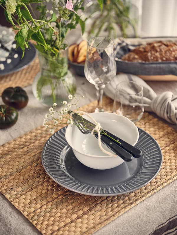 Ein rustikaler Esstisch mit einem grauen Teller, einer weissen Schüssel mit silbernem Besteck, Gläsern und einer Blumenvase auf einer gewebten Tischset.