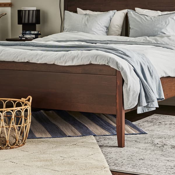 Ein Schlafzimmer mit einem dunkelbraun lasierten HEMNES Bettgestell ist zu sehen, das auf verschiedenen Teppichen steht.