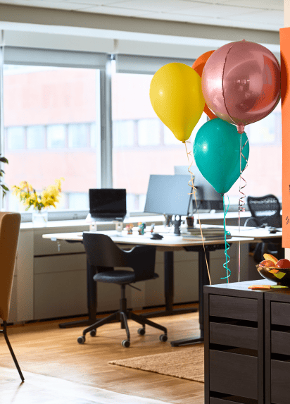 Ein Schreibtisch an einem großen Fenster. Im Vordergrund sind drei Luftballons zu sehen.