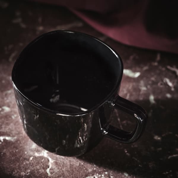 Ein schwarzer glänzender BACKIG Becher mit einem Griff steht auf einer dunkelroten Marmoroberfläche mit etwas Stoff im Hintergrund. 