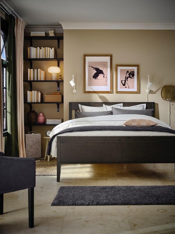 Ein schwarzes Bett mit heller Bettwäsche neben einem eingebauten Regal in einem Schlafzimmer mit hellen Fliessen.