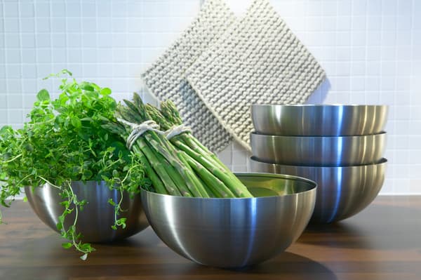 Ein Set von BLANDA Edelstahlschüsseln, in denen frische Kräuter und Spargel liegen, im Hintergrund sind zwei helle Topflappen.