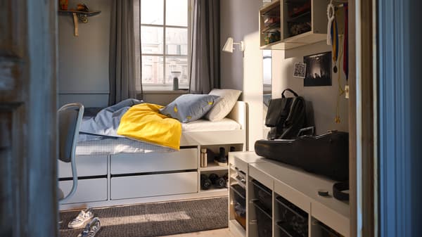 Ein SLÄKT Bettgestell mit blauer Bettwäsche steht in einem kleinen Zimmer vor einem Fenster. An der Wand und auf dem Boden sind TROFAST Aufbewahrungselemente zu sehen.