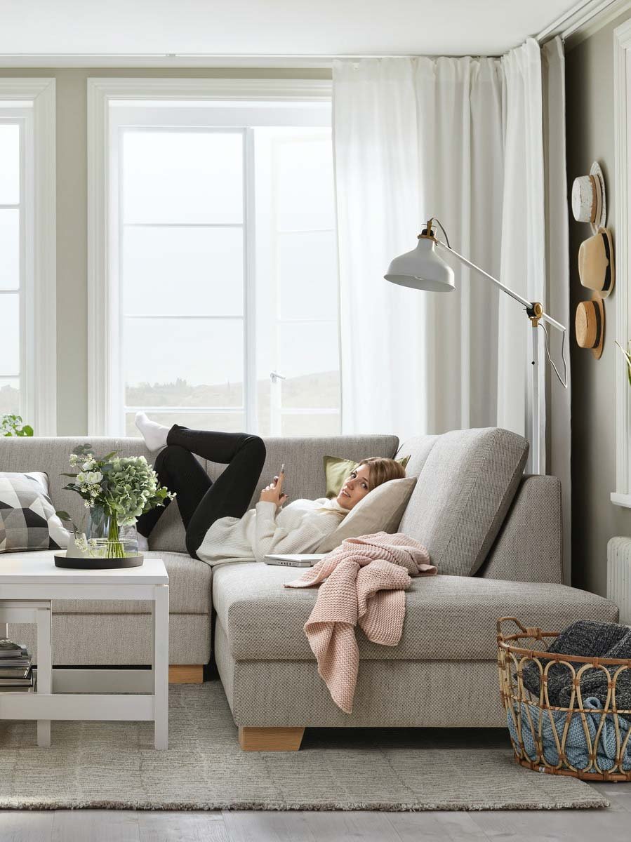 Ein SÖRVALLEN Ecksofa mit offenem Ende steht vor einem Wohnzimmerfenster. Darauf liegt eine junge Frau mit einem Handy in der Hand sie sucht die für sie passende SÖRVALLEN Sofa Variante.