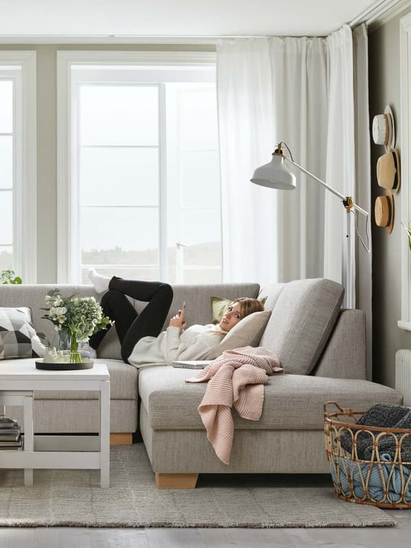 Ein SÖRVALLEN Ecksofa mit offenem Ende steht vor einem Wohnzimmerfenster. Darauf liegt eine junge Frau mit einem Handy in der Hand sie sucht die für sie passende SÖRVALLEN Sofa Variante. 