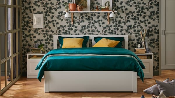 Ein SONGESAND Bettgestell in Weiß mit grüner Bettwäsche und gelben Kissen. Darüber befindet sich ein Regal an der Wand. Über dem Doppelbett hängt eine Hängeleuchte.