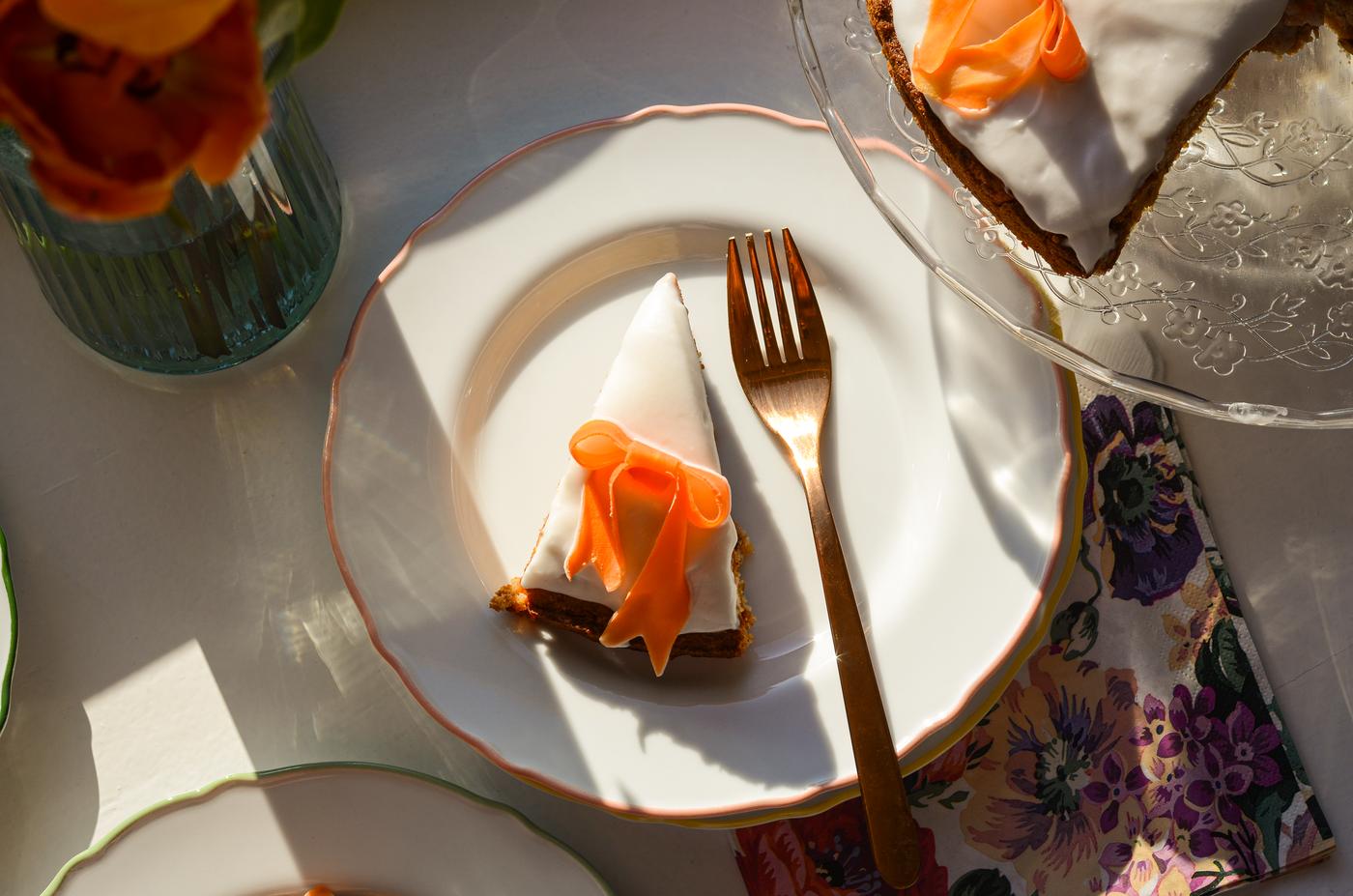 Ein Stück Eistorte mit Orangenblütenverzierung auf einem weissen Teller neben einer goldenen Gabel, daneben eine Vase mit Orangenblüten.