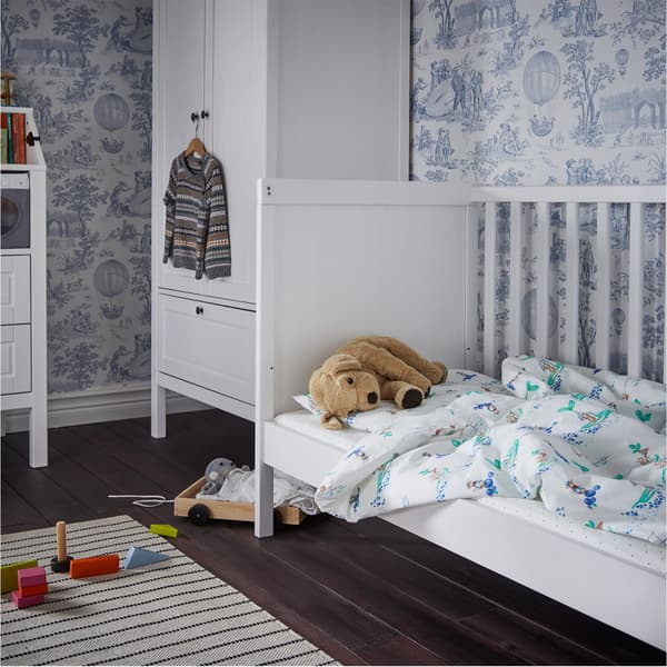 Ein SUNDVIK Babybett in Weiss mit einem Stofftier auf der Matratze und einem Kleiderschrank am Kopfende.