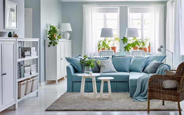 Ein überwiegend in Weiß gehaltenes Wohnzimmer mit Essbereich mit HOLMSUND Eckbettsofa mit Bezug „Orrsta“ hellblau, BYHOLMA Sessel in Grau, KRAGSTA Satztischen in Weiß und mehreren HEMNES Schränken mit 2 Türen weiß gebeizt