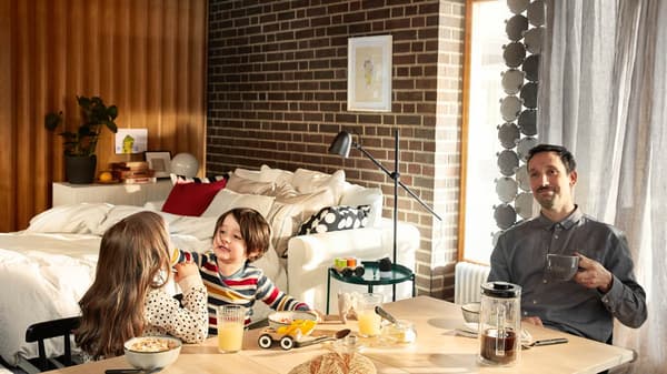 Ein Vater sitzt entspannt am Frühstückstisch und genießt seinen Kaffee, während seine zwei Kinder mit dem Essen spielen.