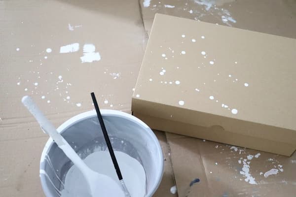 Ein VATTENTRÅG Kasten aus Kraftpapier liegt auf einer Pappunterlage. Daneben ein Becher mit weißer Farbe darin ein Pinsel. 