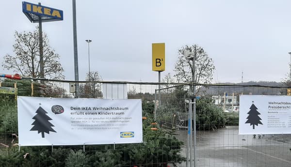 Ein Weihnachtsbaumverkauf auf dem Parkplatz eines IKEA-Marktes. Im Vordergrund sind zwei große Banner befestigt, eines mit der Aufschrift „Dein IKEA Weihnachtsbaum erfüllt einen Kindertraum“ und eines mit einer Preisübersicht für verschiedene Baumarten. Dahinter sind zahlreiche aufgestapelte Weihnachtsbäume zu sehen. Im Hintergrund ragt das große IKEA-Schild in den grauen Himmel.  