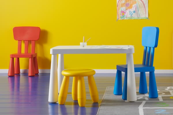 Ein weißer Kindertisch mit einem gelben Hocker, einem blauen und einem roten Stuhl aus der MAMMUT Kindermöbel Serie.