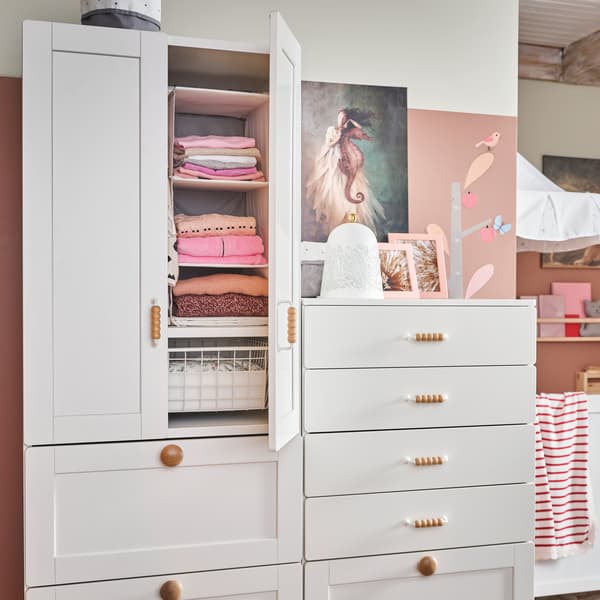Ein weißer Kleiderschrank mit drei Schubladen und zwei Türen mit Holzgriffen. Eine Tür steht offen und gibt den Blick auf gefaltete Kleidung in Aufbewahrungsfächern frei.