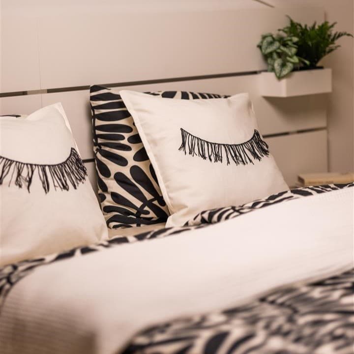 Ein weißes Bett steht an einer Wand das weiß, schwarze Bettwäsche hat und Pölster mit einem Augen Muster.